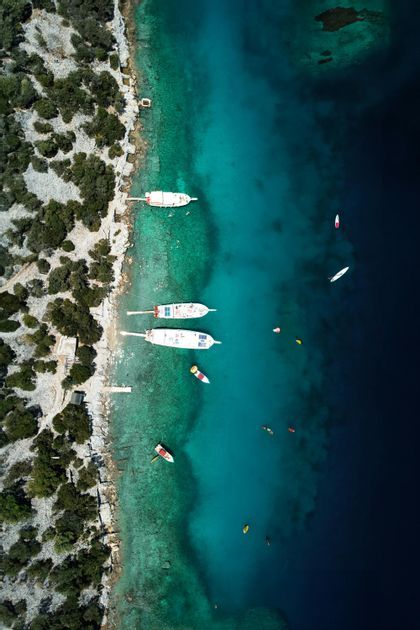 Vista aerea di imbarcazioni e kayak lungo una costa rocciosa con acqua cristallina turchese e blu profondo.