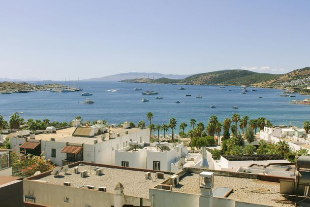 Vista dall'alto di candidi edifici costieri che si affacciano su una placida baia blu, costellata di numerosi yacht e imbarcazioni, sotto un cielo sereno.