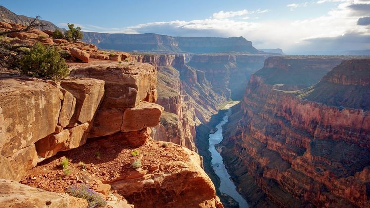 Depuis une falaise de roche rouge, une rivière serpente à travers un canyon profond et vaste, sous un ciel bleu parsemé de nuages.