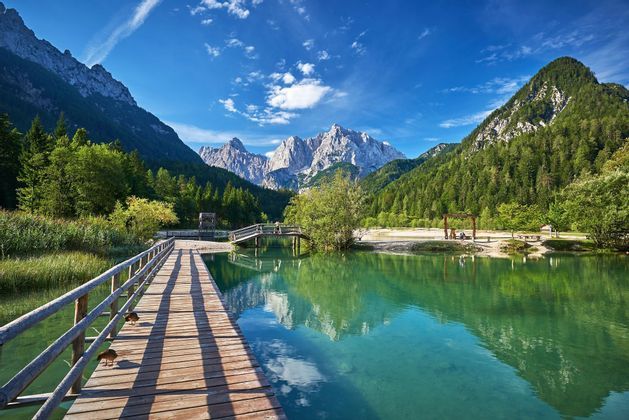 Ein Holzsteg führt über einen ruhigen, türkisen See zu bewaldeten Hügeln und schroffen Berggipfeln unter blauem Himmel.