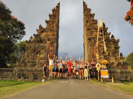 Un groupe WeRoad en voyage saute joyeusement pour une photo devant une grande et ancienne porte de pierre fendue.