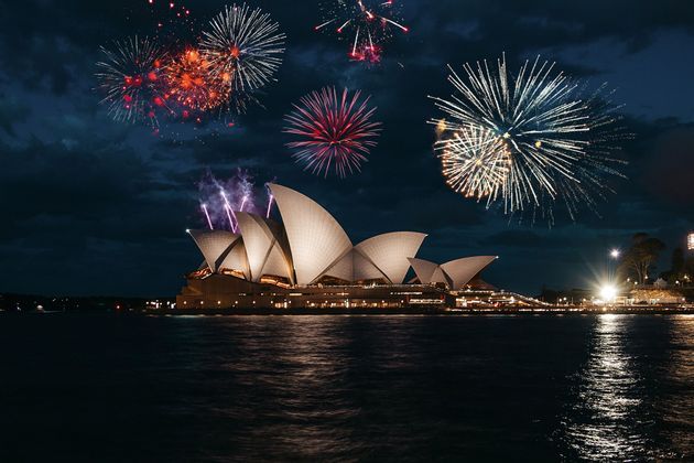 Colorful fireworks explode in the night sky above the illuminated Sydney Opera House, viewed from across the water.