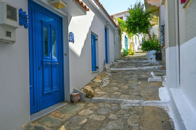 Una stretta via acciottolata si snoda in salita tra edifici imbiancati a calce con porte e persiane di un blu vivace.