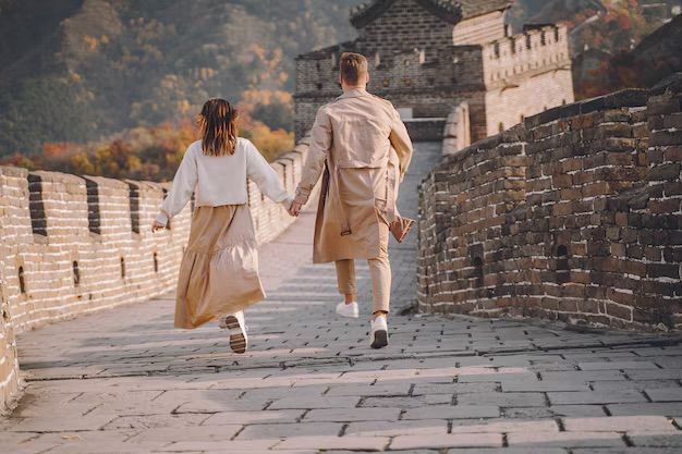 Una pareja vista de espaldas, tomados de la mano y corriendo por la cima de una gran muralla de piedra histórica con montañas al fondo.