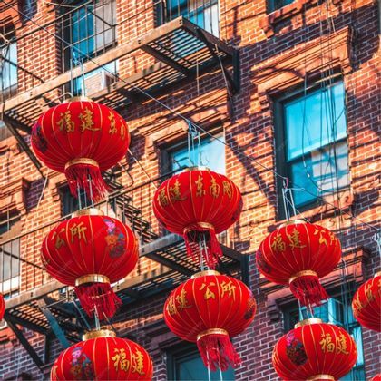 Farolillos chinos rojos con letras doradas cuelgan de cuerdas frente a un edificio de ladrillo rojo con escalera de incendios.