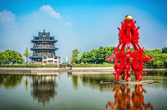 Una pagoda china tradicional y una escultura de dragón rojo reflejadas en un lago en calma bajo un cielo azul brillante.