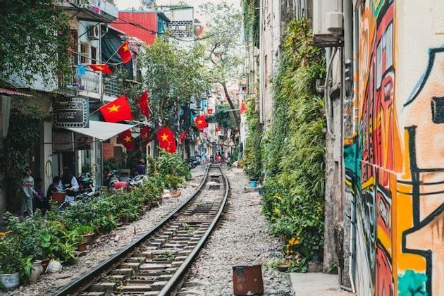 Eine einzelne Bahntrasse verläuft durch eine schmale Gasse, die von Gebäuden, vietnamesischen Flaggen, Topfpflanzen und graffiti-bedeckten Wänden gesäumt wird.