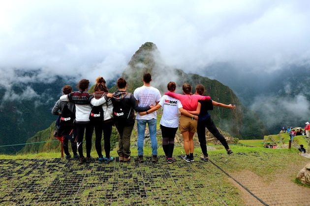 Eine WeRoad-Gruppe steht Arm in Arm und blickt von einem grasbewachsenen Aussichtspunkt auf einen wolkenverhangenen Berggipfel.