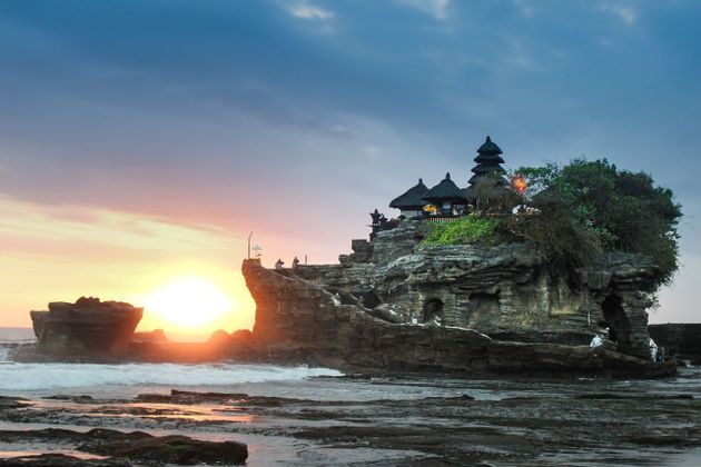 Un templo con techos escalonados se alza en una isla rocosa en el mar al atardecer, con las olas lamiendo la orilla.