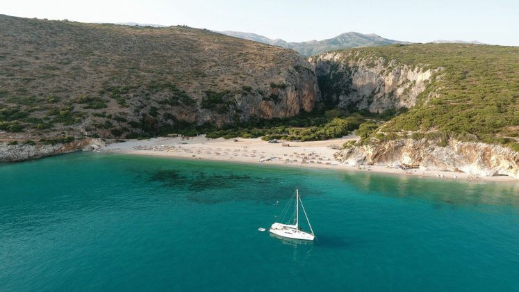 An aerial view of a white sailboat floating on turquoise water in a cove with a sandy beach and steep, green cliffs.