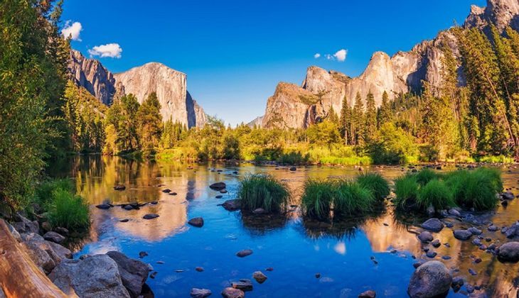 A calm river reflects towering granite cliffs and a dense forest under a bright blue sky.