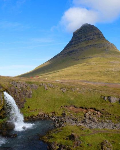 Una pequeña cascada que cae a una poza en primer plano, con una gran montaña verde cónica al fondo bajo un cielo azul.