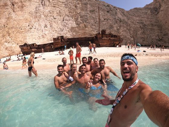 Un homme prend un selfie avec son groupe WeRoad sur une plage aux eaux peu profondes, avec une grande épave rouillée en arrière-plan.