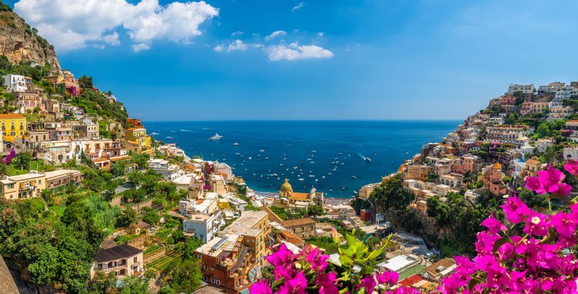 Un colorato borgo arroccato su una ripida scogliera si affaccia su un mare blu con molte barche, incorniciato da fiori rosa.