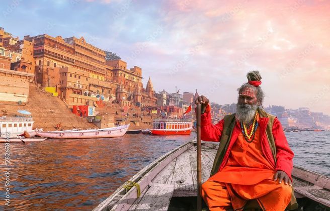 A man with a long grey beard and orange robes sits at the front of a wooden boat on a river, with city buildings along the waterfront.