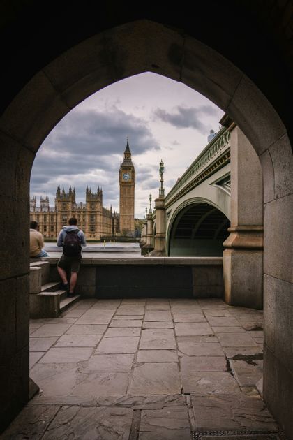 Due persone ammirano la torre dell'orologio del Big Ben e il Palazzo di Westminster da sotto un arco di pietra in una giornata nuvolosa.