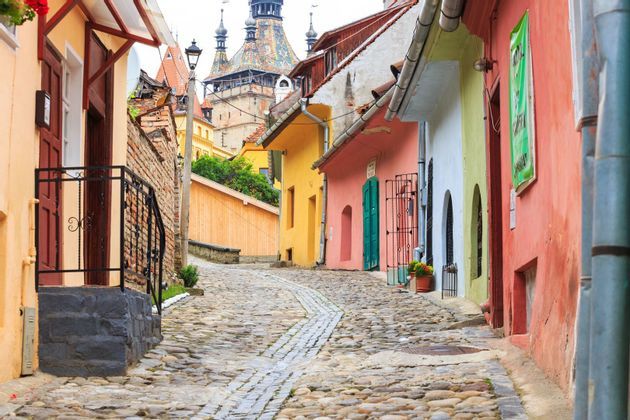 Una calle empedrada, estrecha y vacía que serpentea cuesta arriba pasando por edificios coloridos hacia una torre de reloj en un pueblo histórico.