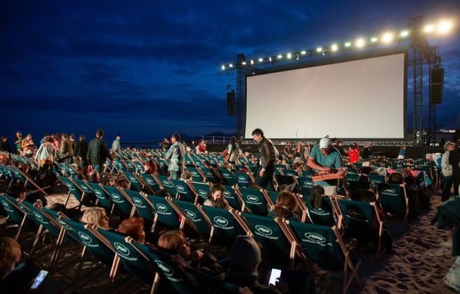 El público se sienta en filas de tumbonas verdes en una playa por la noche, frente a una gran pantalla de cine al aire libre iluminada.
