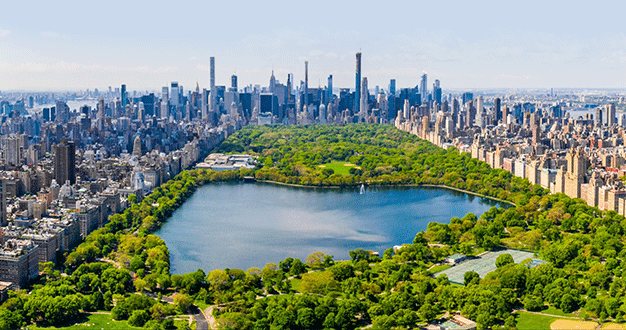Una vista aérea de un gran parque urbano con un lago, enclavado entre densas manzanas de la ciudad llenas de rascacielos en un día soleado.