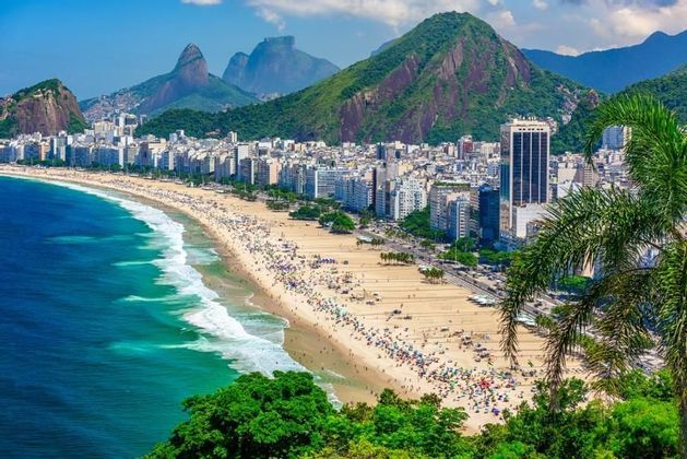 Vue aérienne d'une plage de sable bondée s'étendant le long d'un paysage urbain dense, avec des montagnes vertes en arrière-plan sous un ciel bleu.