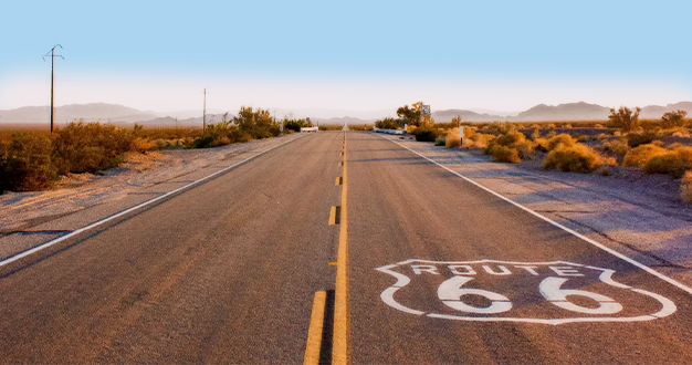Le logo du bouclier de la Route 66 peint sur une route asphaltée qui s'étend à travers un paysage désertique vers des montagnes lointaines.