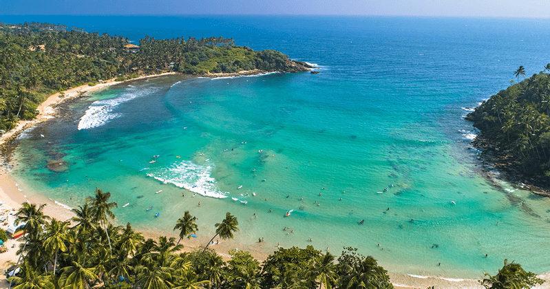 Vista aerea di una baia tropicale dove le persone fanno surf e nuotano in acque turchesi, accanto a una spiaggia sabbiosa orlata di palme.