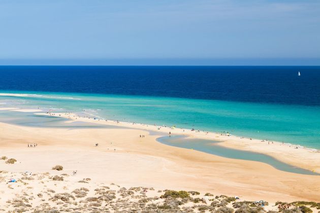 Une vue en plongée d'une plage de sable où des personnes sont dispersées le long du rivage turquoise, avec l'océan d'un bleu profond en arrière-plan.