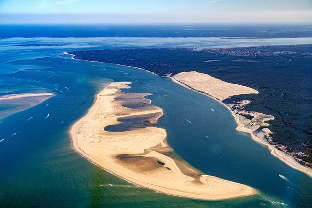 Veduta aerea di una grande duna e banchi di sabbia lungo una costa ricoperta da una fitta foresta verde, con barche in acqua.