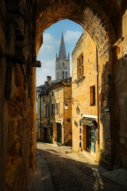 Una vista attraverso un arco in pietra lungo una stretta via acciottolata verso il campanile di una chiesa sotto un cielo blu.