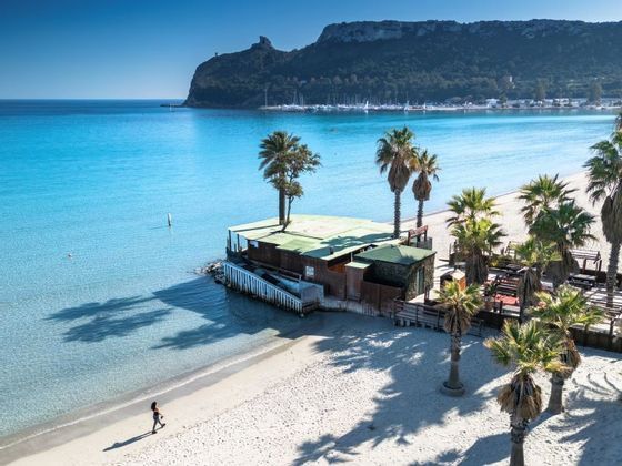 Una vista aerea di una persona che cammina su una spiaggia di sabbia bianca, bagnata da acque cristalline color turchese, con palme e un promontorio.