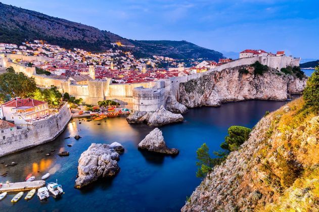Une ville historique fortifiée sur la côte est illuminée au crépuscule, surplombant une baie calme avec de petits bateaux.