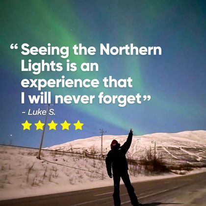 A person in winter clothes stands on a snowy road making a peace sign under the green glow of the Northern Lights at night.