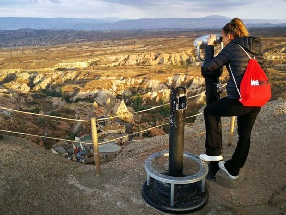 Una donna con uno zaino WeRoad rosso guarda attraverso un mirino, ammirando una vasta valle rocciosa da un punto panoramico elevato.