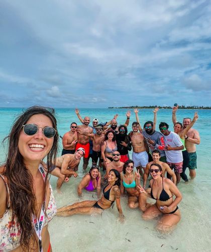 A woman takes a selfie with a WeRoad group trip as they pose and smile in shallow, clear turquoise water under a cloudy sky.