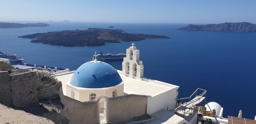 Una chiesa bianca con una cupola blu e un campanile domina un mare blu profondo con isole vulcaniche e navi da crociera in lontananza.