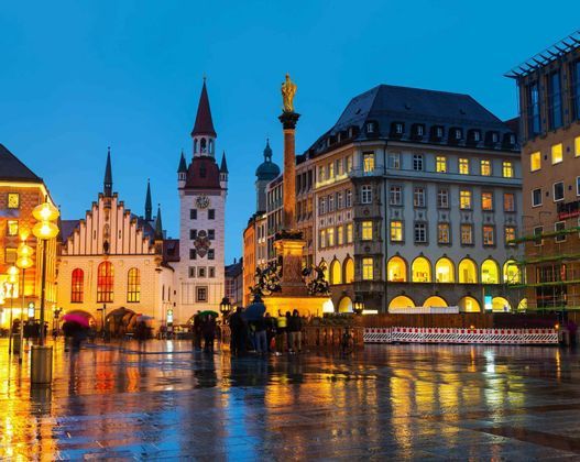 A historic city square with a clock tower and a column, illuminated at dusk with lights reflecting on the wet cobblestone ground.