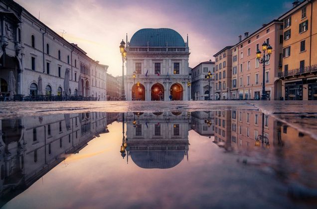 Una toma contrapicada de un edificio histórico con cúpula, su reflejo capturado en un charco en una plaza de la ciudad al atardecer.