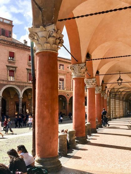 Una fila de columnas de terracota con capiteles ornamentados bordea un pórtico, con gente visible en la soleada plaza al fondo.