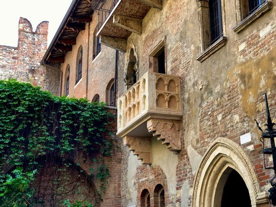 Un balcone in pietra finemente lavorato sporge dal muro di mattoni antichi di un edificio storico, con edera rigogliosa nelle vicinanze.
