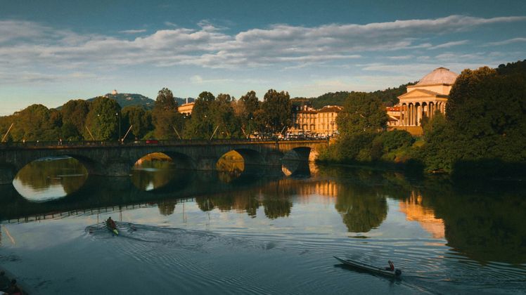 Un puente de arco de piedra sobre un río tranquilo con dos barcos, reflejando los edificios de la ciudad y los árboles al atardecer.