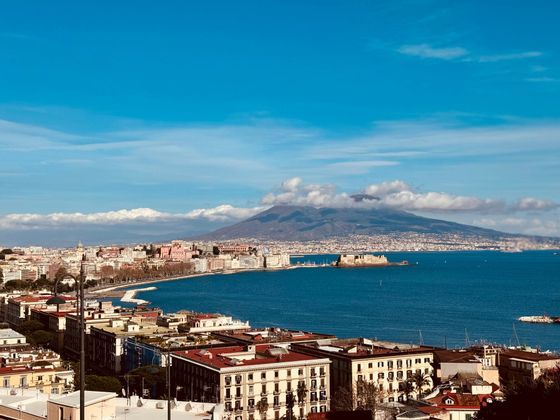 Panorámica de una ciudad costera a orillas de una bahía azul, con un volcán imponente al fondo bajo un cielo claro.