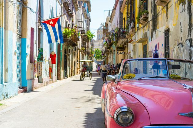 Ein pinkfarbenes Oldtimer-Cabrio parkt auf einer sonnigen Stadtstraße, gesäumt von farbenfrohen Gebäuden, von denen an einem eine kubanische Flagge hängt.
