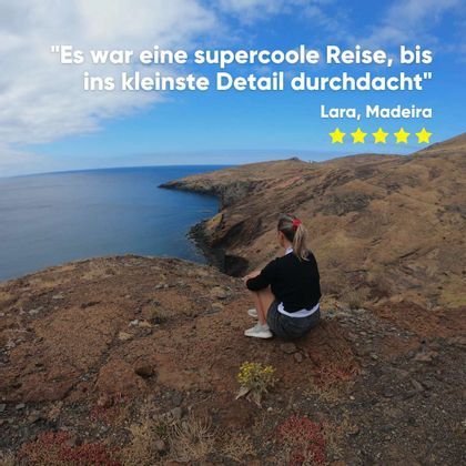 Eine blonde Frau sitzt auf einer felsigen Klippe und blickt auf das Meer und eine zerklüftete Küstenlandschaft unter einem teilweise bewölkten Himmel.