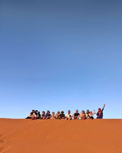 A WeRoad group trip sits together on the crest of a vast orange sand dune under a clear blue sky.
