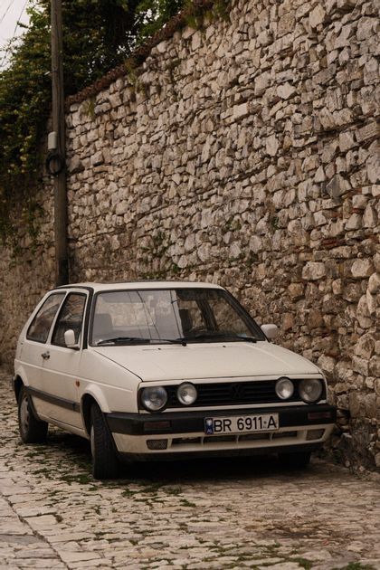 Une voiture à hayon vintage blanche est garée dans une étroite rue pavée contre un haut mur de pierre.