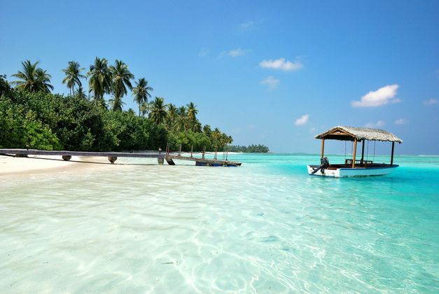 Ein kleines Boot mit Reetdach, vertäut in seichtem türkisfarbenem Wasser neben einem Sandstrand mit Palmen und einem Holzsteg.