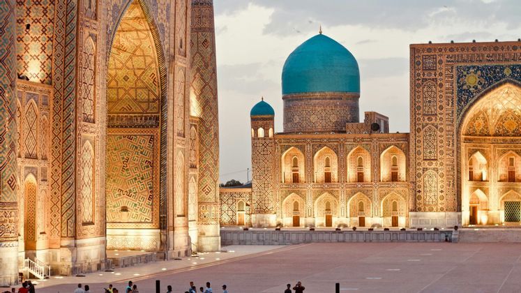 Edifici riccamente ornati e illuminati, con cupole blu e intricate maioliche, si affacciano su un'ampia piazza al tramonto.