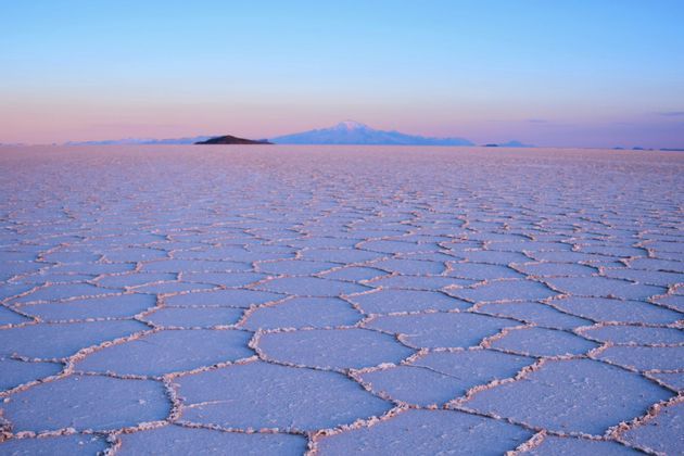 A vast salt flat with hexagonal patterns stretches towards distant mountains under a colorful sky at sunrise.
