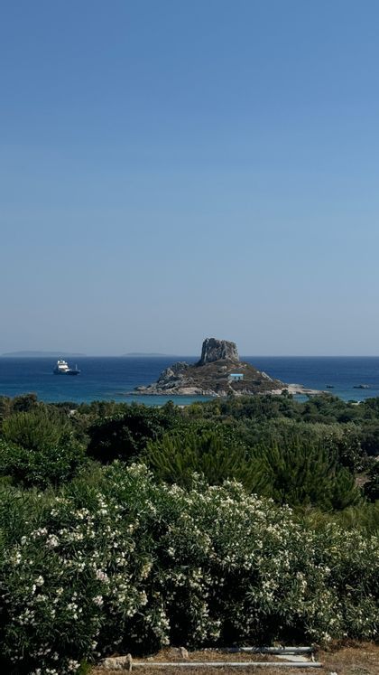 Una vista suggestiva di un isolotto roccioso con un piccolo edificio dal tetto blu in mezzo al mare, incorniciata da cespugli verdi, mentre un traghetto passa sotto un cielo sereno.