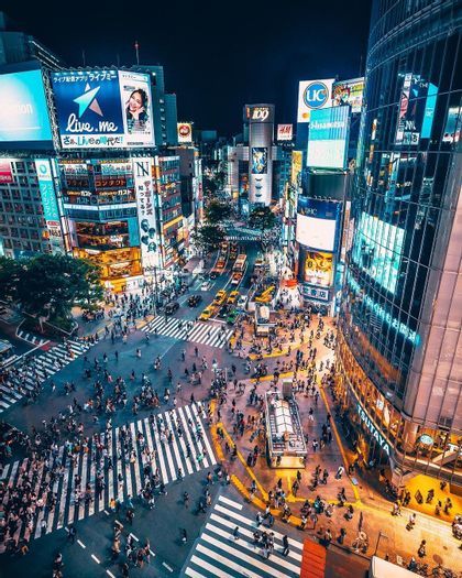 An aerial view of a bustling city intersection at night, with crowds of people and cars crossing under bright neon signs.